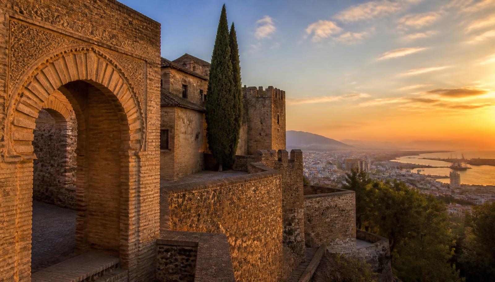 Murallas y arcos de herradura de la Alcazaba de Málaga al atardecer con la ciudad y el puerto al fondo